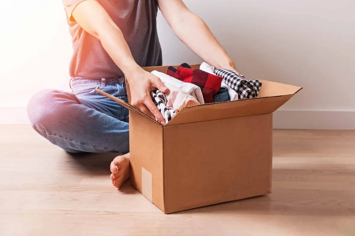 person packing folded clothes into cardboard box sitting on floor at home
