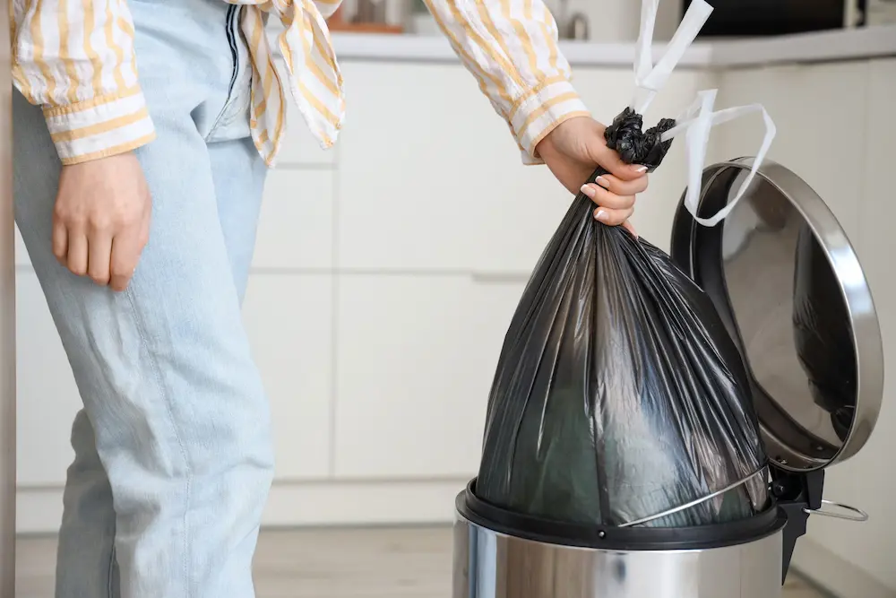 A woman throwing a black garbage bag into a stainless steel trash bin, emphasizing the importance of proper garbage disposal to prevent pest infestations.